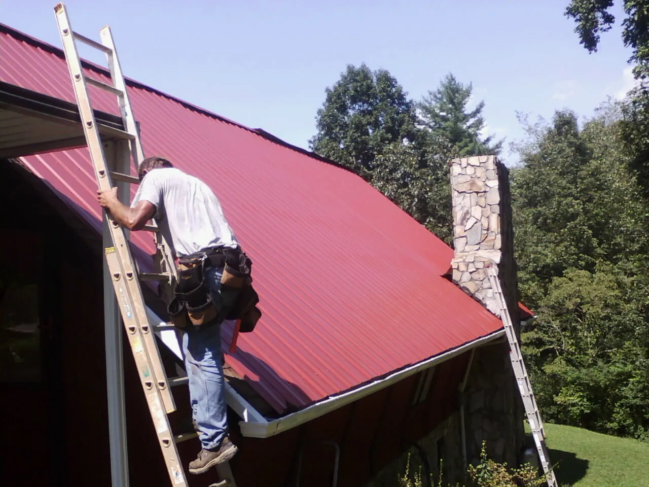 Roofer on a ladder beside a red metal roof and stone chimney — Soffit & Fascia Repair by Roofing Brothers in Mappsville